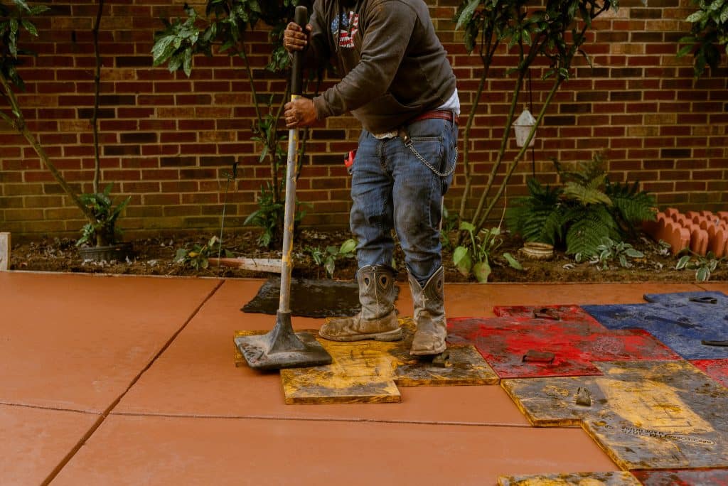 A construction worker in boots and jeans uses a tamper tool to press colorful concrete stamps onto a freshly poured orange slab, showcasing the skill of a concrete contractor, with a brick wall and plants in the background.