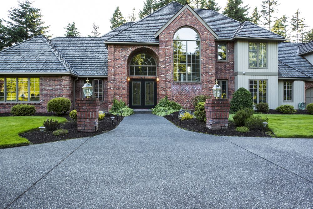 A large two-story brick house with a steep roof, large arched windows, and double front doors. The paved driveway, expertly crafted by a concrete contractor, is lined with manicured lawns, shrubs, and lanterns on brick pillars at the entrance.