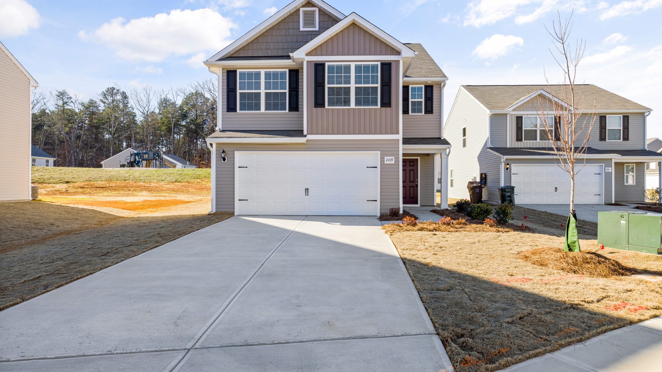 A two-story suburban house with tan siding, black shutters, and a white double garage. The driveway is wide and clean, and the yard has sparse grass. Neighboring houses and trees are visible in the background.