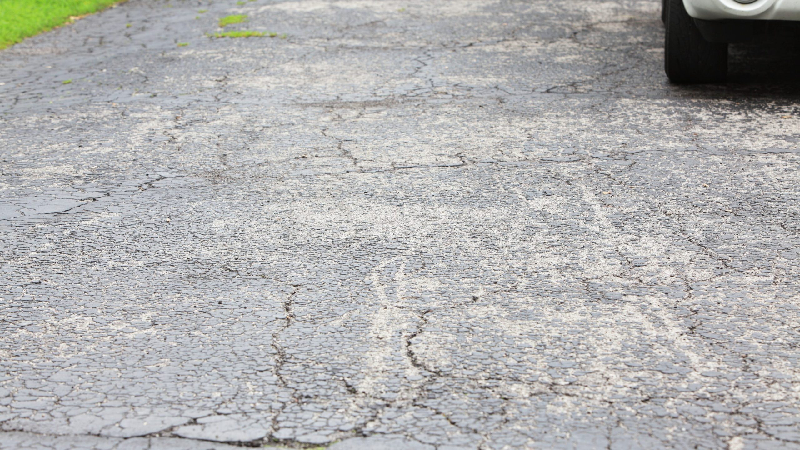 A close-up view of a cracked asphalt driveway with visible rough patches hints at reduced driveway lifespan; part of a vehicle is seen at the top right corner in Greenville SC.