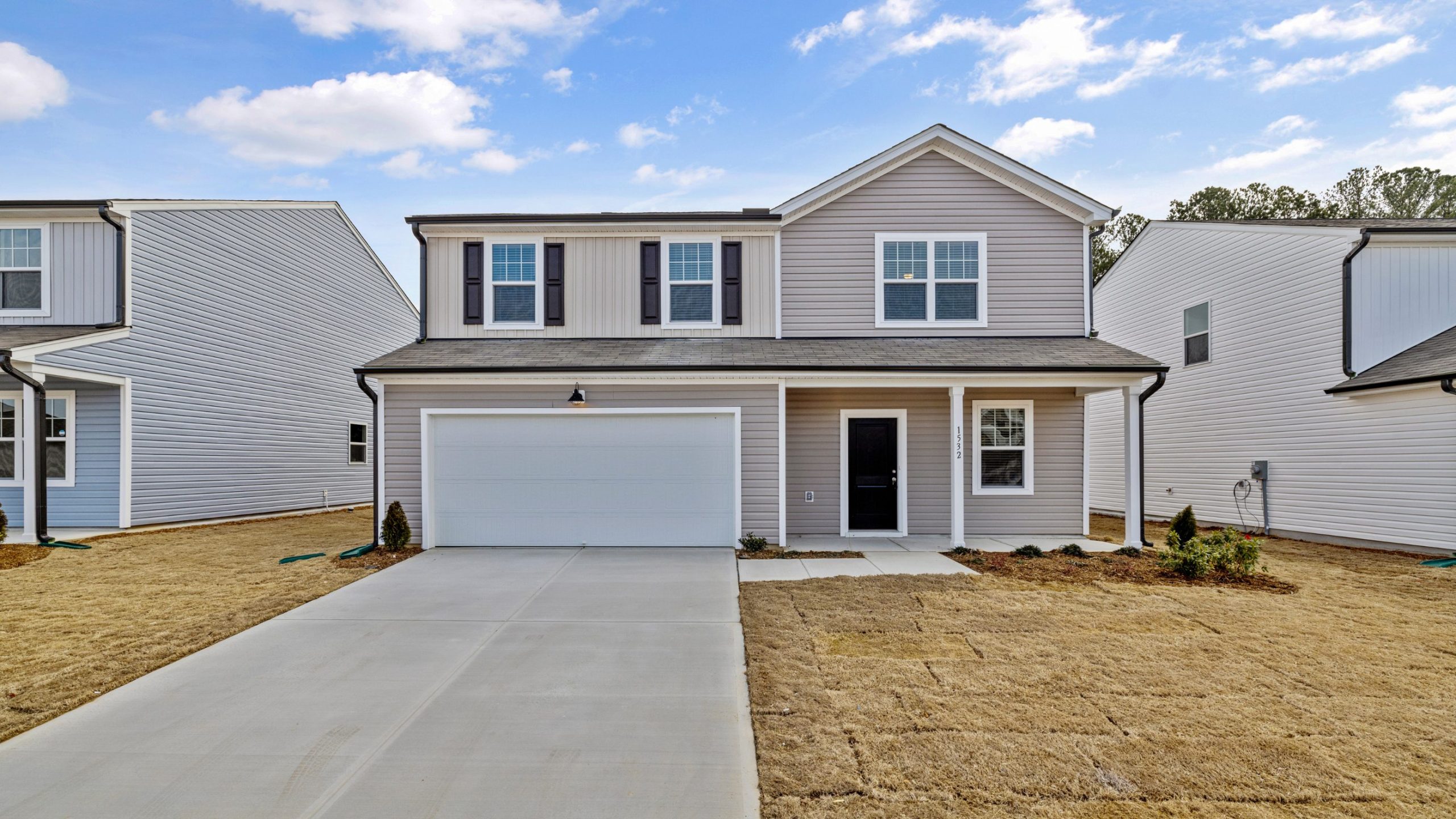 A two-story suburban house with gray siding, white trim, a double garage, and a concrete driveway. Small landscaped areas with shrubs flank the entrance, and the lawn is dry and brown. Blue sky with scattered clouds overhead.