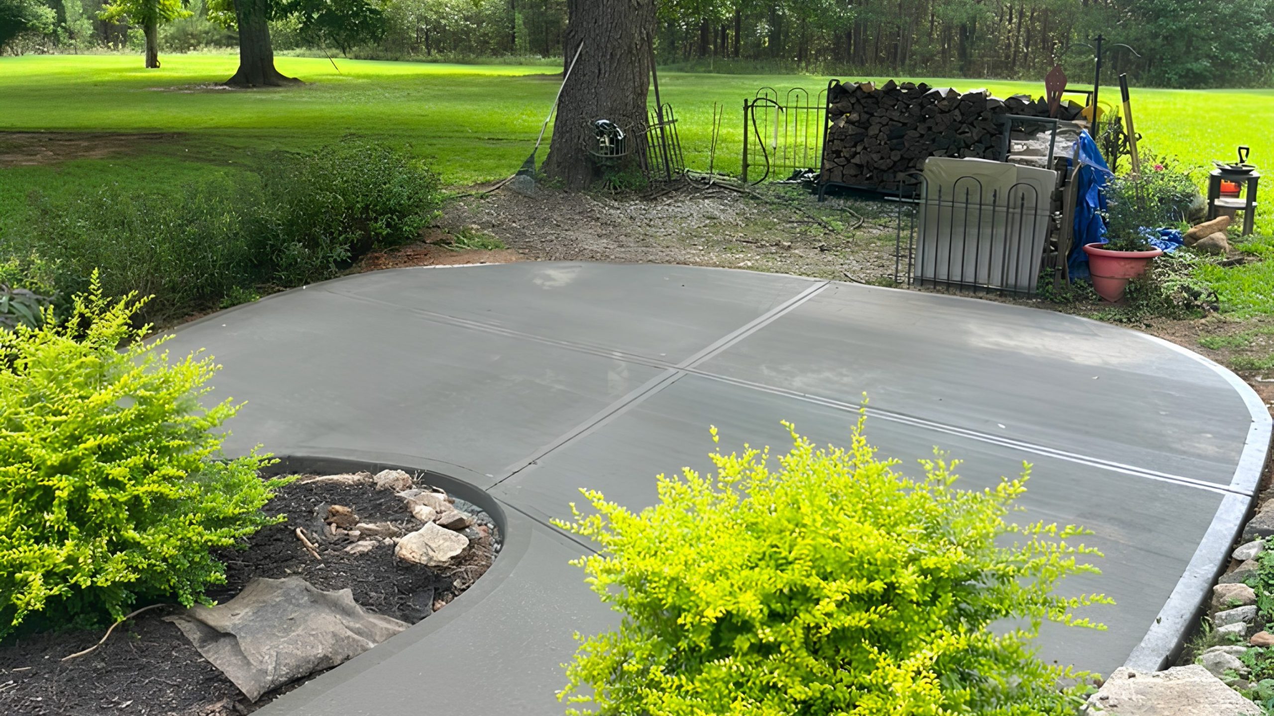 A freshly poured, smooth concrete patio curves around landscaped bushes, with stacked firewood and tools organized near a tree in a grassy backyard.