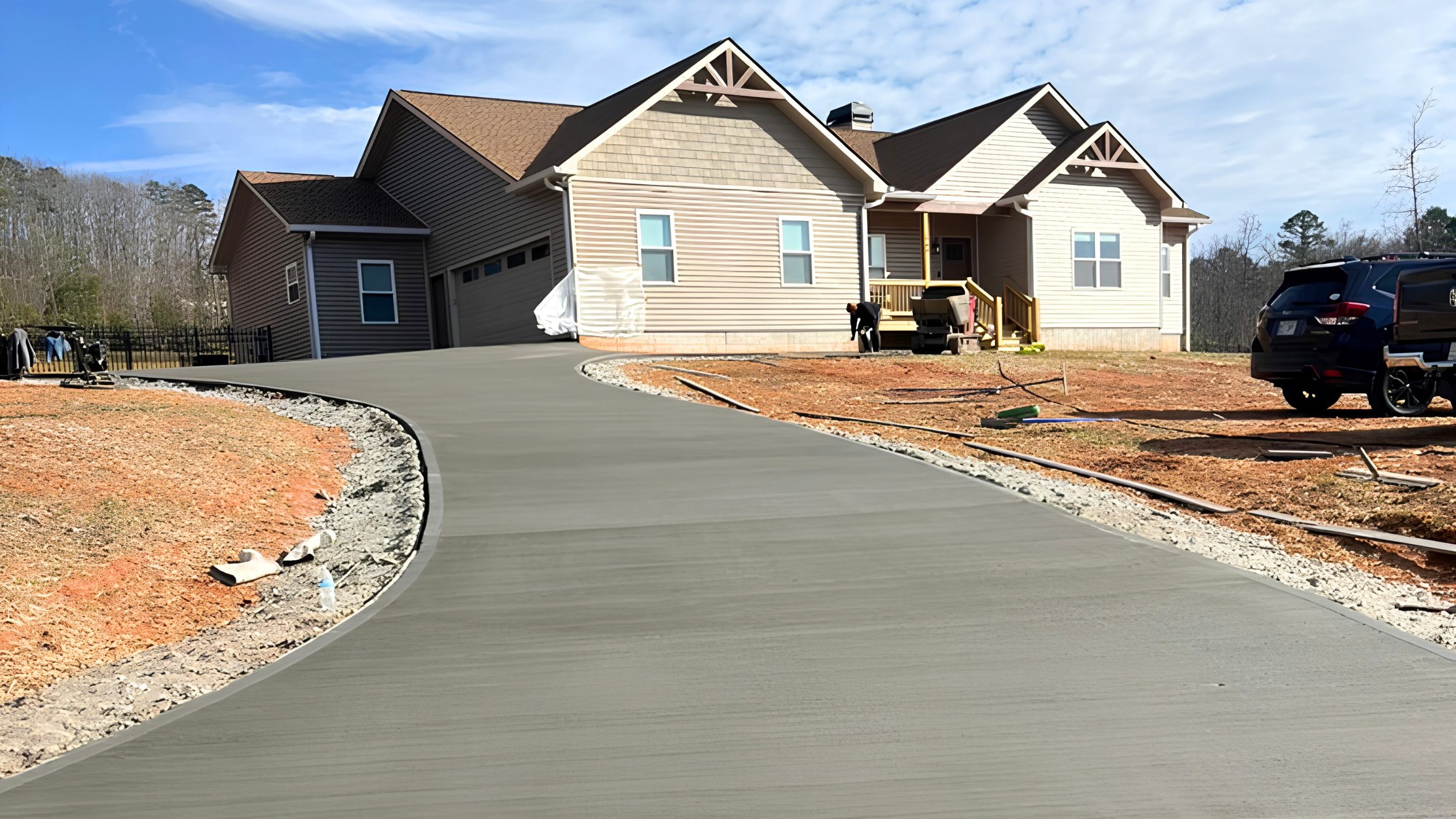 A newly poured concrete driveway, completed after securing a concrete permit Greenville SC requires, curves up to a beige, modern house with a front porch. The yard is unfinished with red soil, and there are a few cars and people near the house.