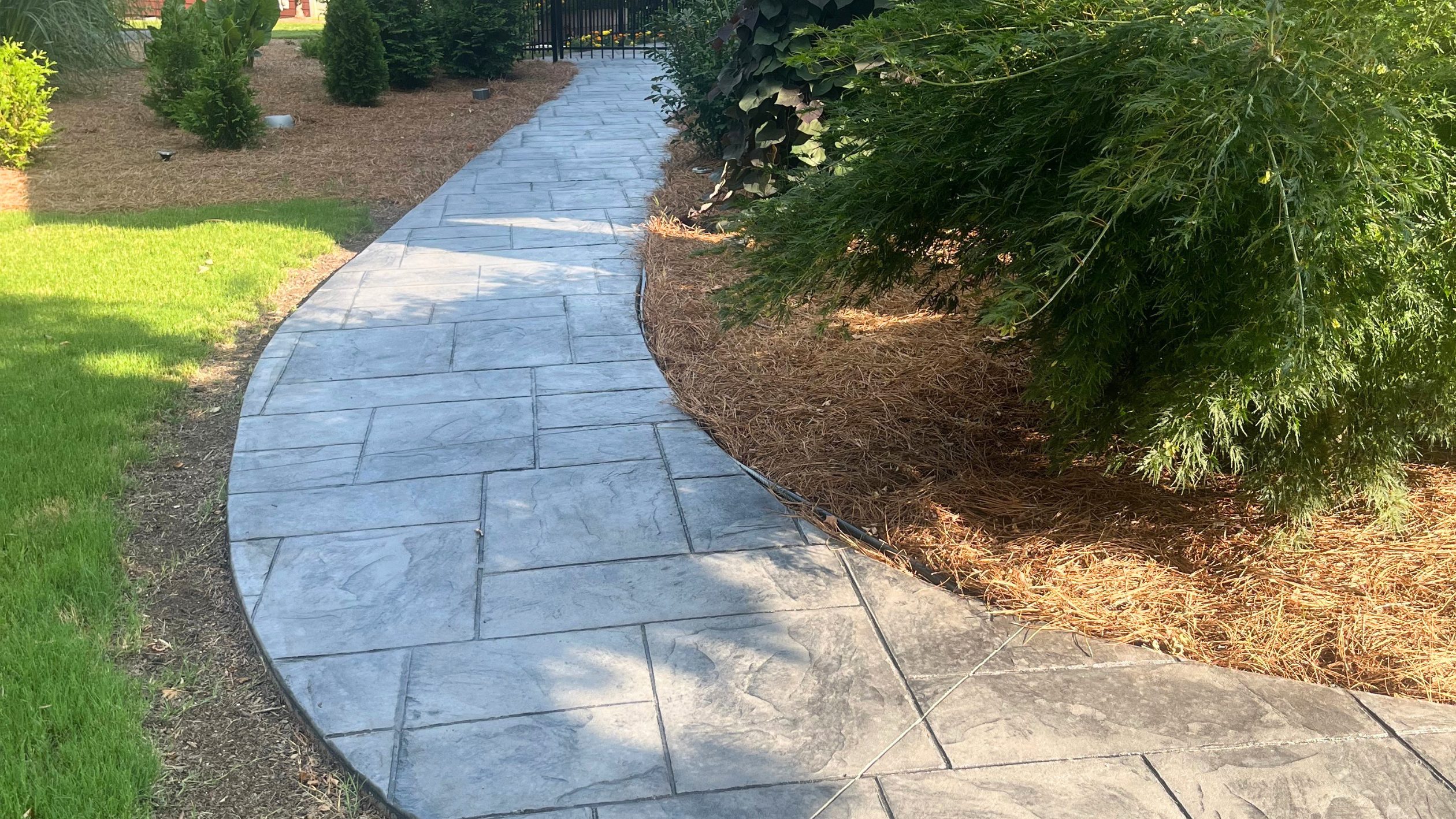 A curved, stamped concrete walkway bordered by green grass on one side and pine straw mulch with bushes on the other, leading toward a gate—perfectly designed for Greenville SC homes and expertly installed with the proper concrete permit in place.