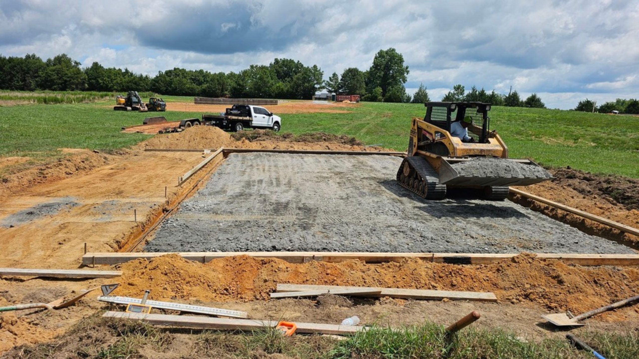 A bulldozer spreads gravel inside a rectangular foundation frame on a construction site, preparing the area for future concrete walkways. In the background, workers, vehicles, and grassy fields can be seen under a partly cloudy sky, reflecting thoughtful concrete design often found in sidewalks throughout Greenville SC.